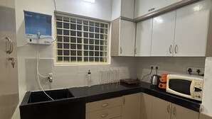 A kitchen with a black countertop, white cabinets, a window with a grid pattern, and a water dispenser. - The golden nest (Hyderabad)