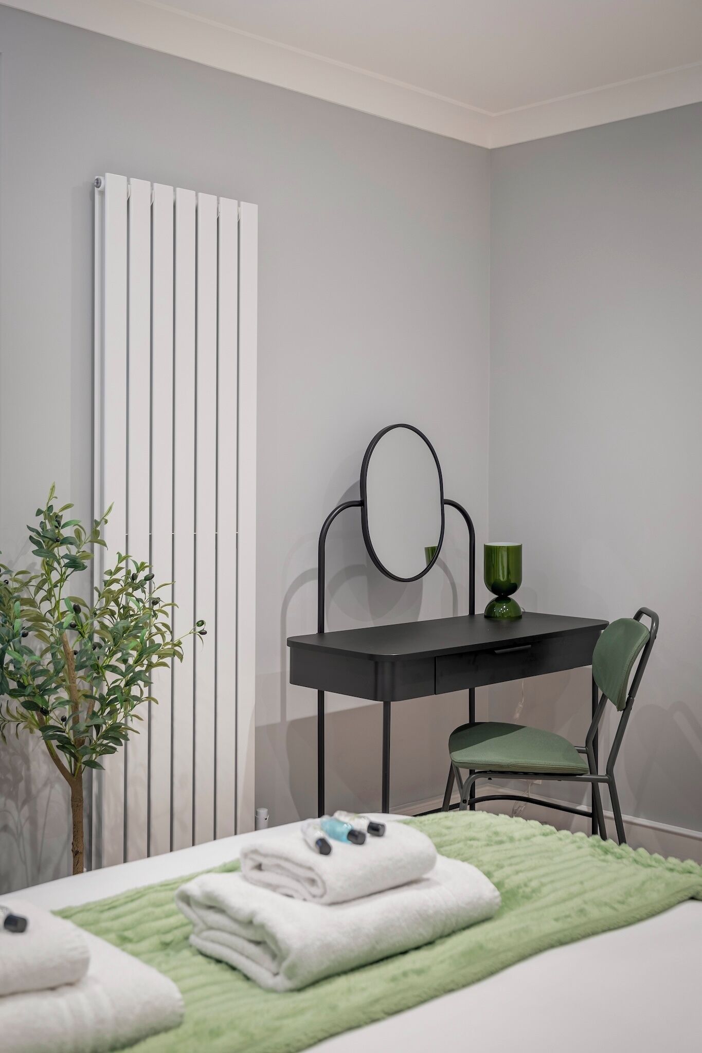 A bedroom with a bed, a green blanket, a bedside table with a mirror, a green chair, a radiator, and a potted plant.