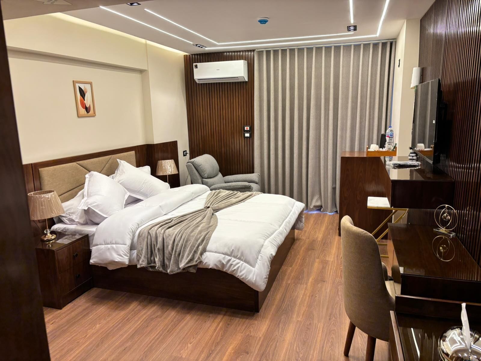 Una habitación de hotel con cama, mesita de noche, lámpara, silla, escritorio, televisor y cortinas.
