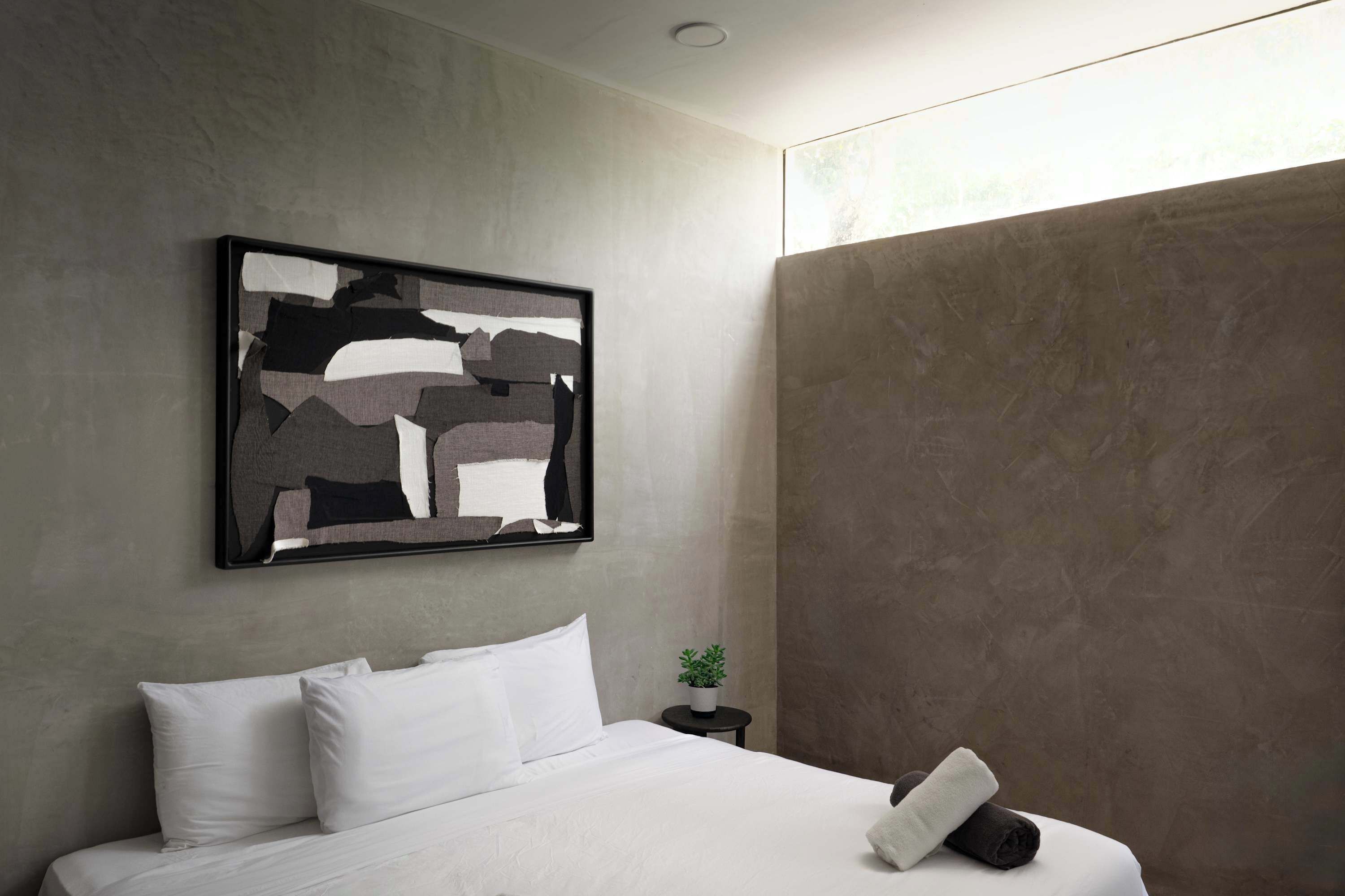 A modern hotel room with a large abstract painting, a neatly made bed, a small potted plant, and a large window.