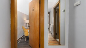 Ein Flur mit einer Holztür, die mit '5' beschriftet ist, einem Spiegel an der Wand, einem gelben Stuhl und einem Schreibtisch mit einer Lampe.