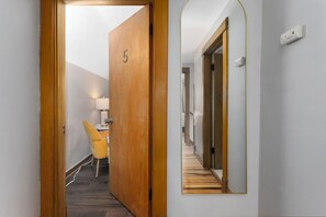 Ein Flur mit einer Holztür, die mit '5' beschriftet ist, einem Spiegel an der Wand, einem gelben Stuhl und einem Schreibtisch mit einer Lampe.