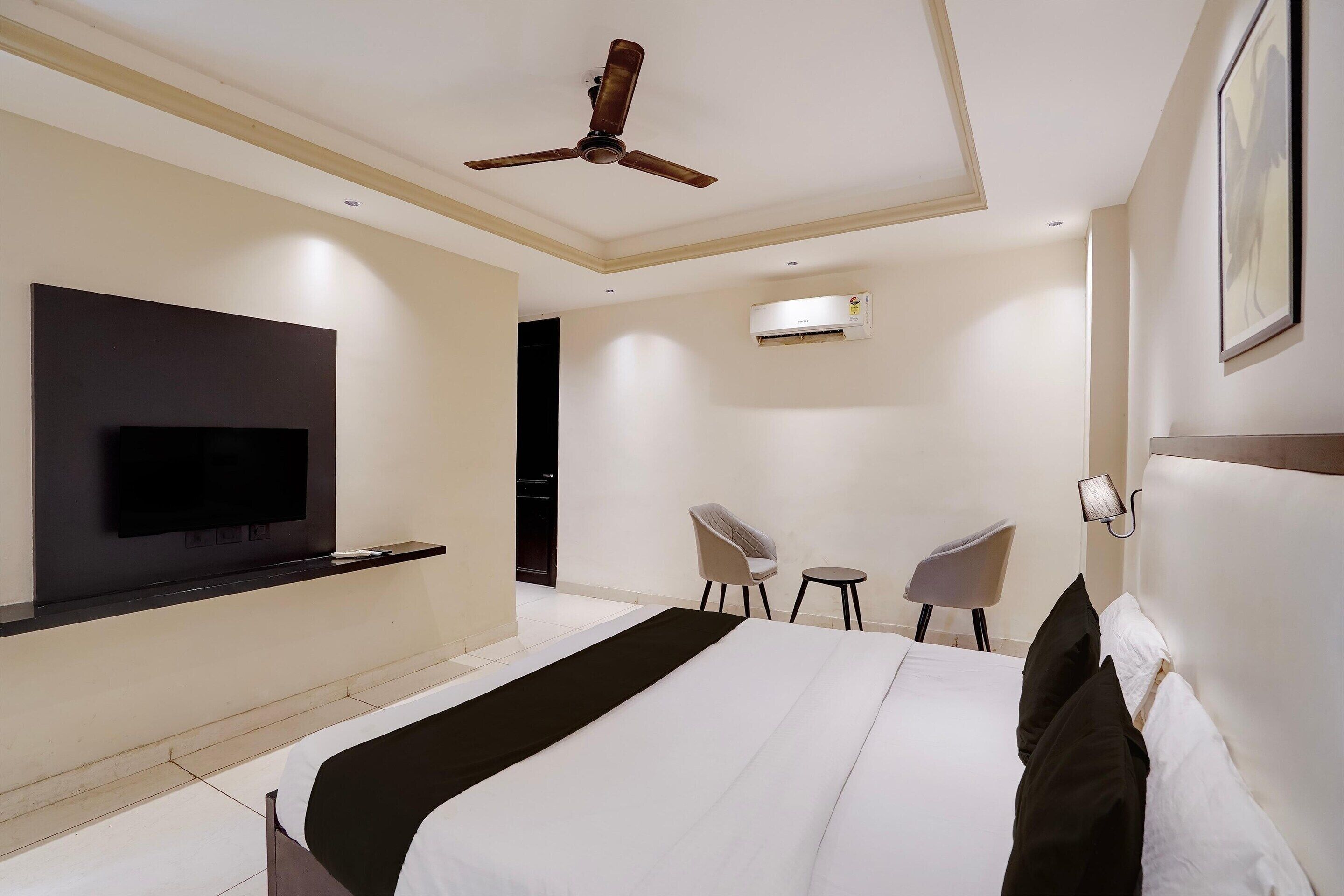 A modern hotel room with a large bed, a flat-screen TV mounted on the wall, a ceiling fan, an air conditioning unit, and two chairs.