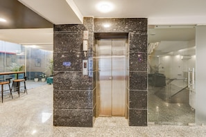 An elevator in a lobby with a marble wall, a glass door, and a seating area with stools. - Deluxecloud Bay (Gurugram)