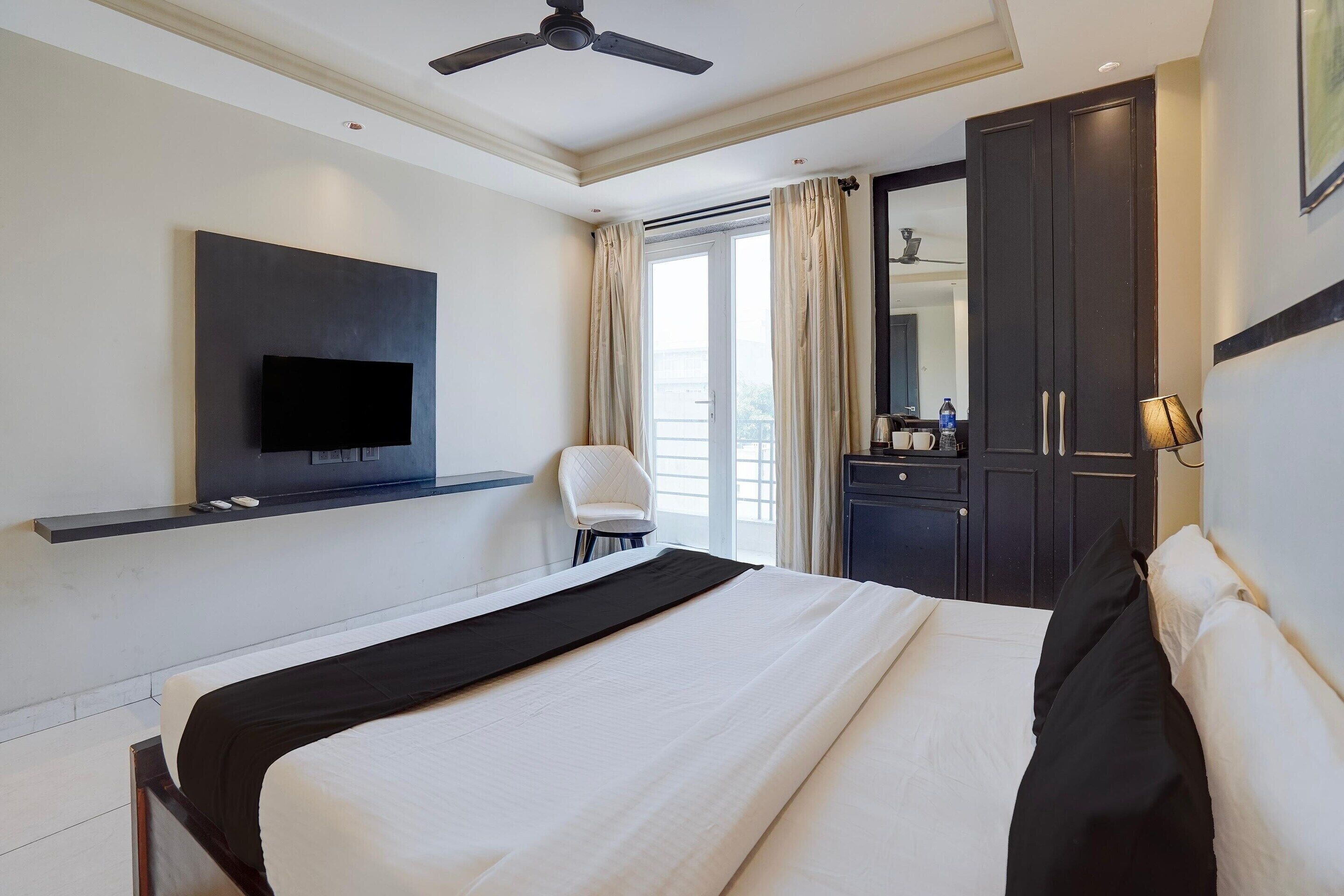 A hotel room with a bed, a television mounted on the wall, a chair, a desk with a water bottle, and a wardrobe.