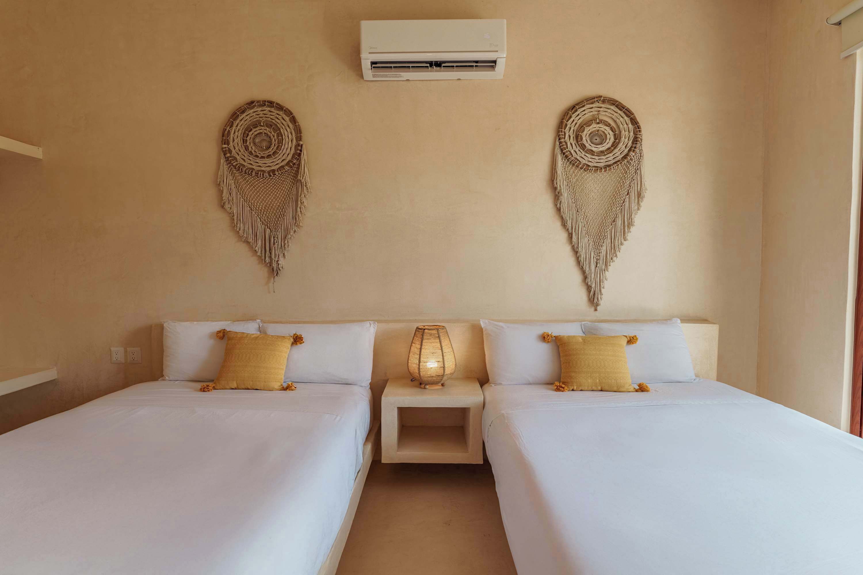 A room with two beds, a bedside table with a lamp, an air conditioning unit, and wall decor.