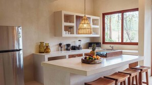 A modern kitchen with a central island, bar stools, a refrigerator, and a window with a view of trees. - Modern ground-level 3BR Retreat with Private Pool (Tulum)