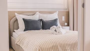 Una cama bien hecha con una manta texturizada y toallas enrolladas, un cabecero de madera y una mesita de noche con un reloj pequeño.
