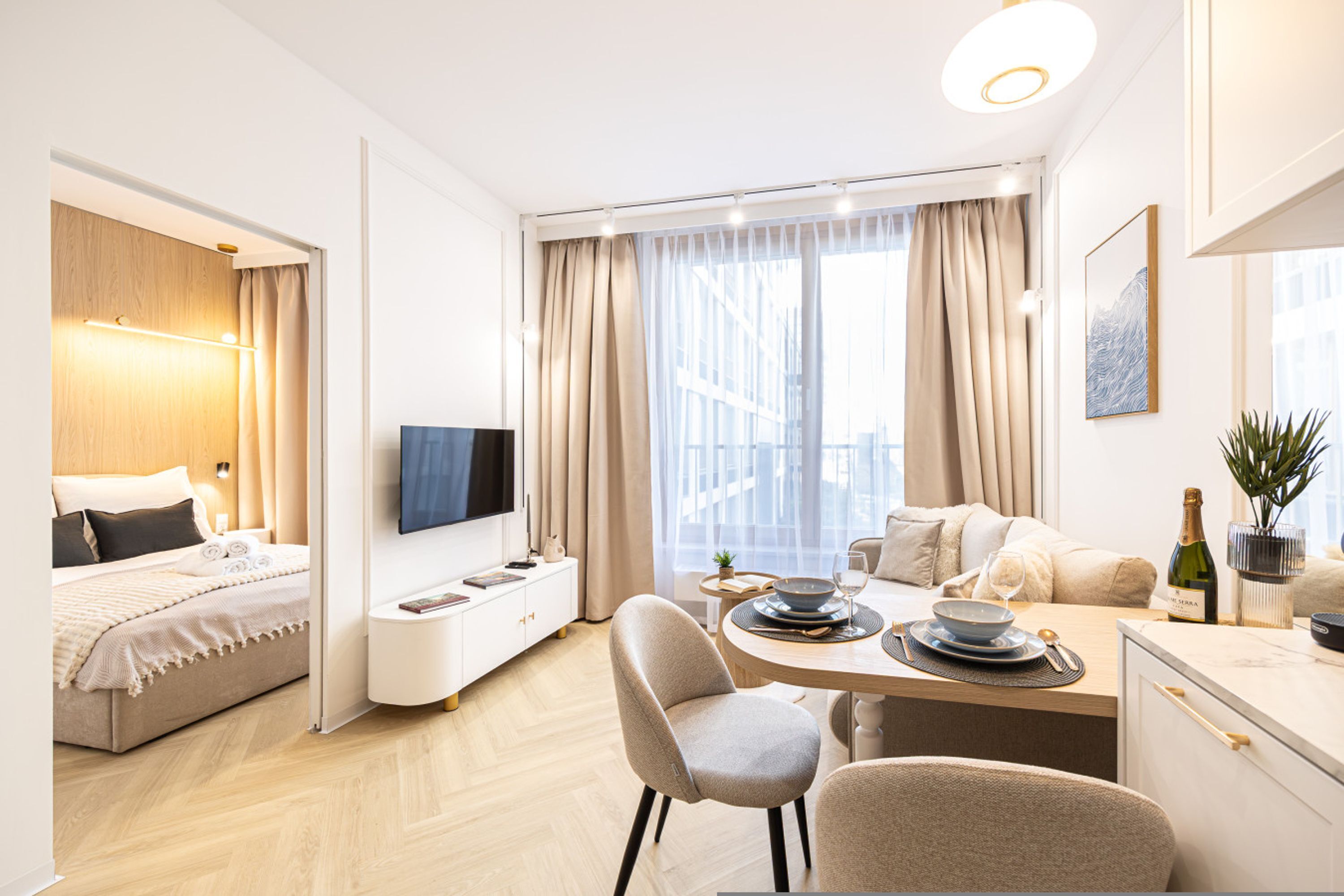 Habitación de hotel moderna con cama, televisor, zona de comedor con mesa preparada para dos personas y vistas panorámicas de la ciudad a través de grandes ventanales.