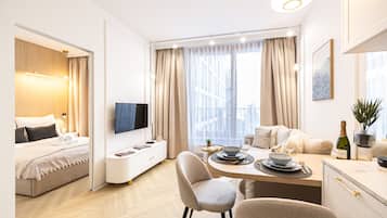 Habitación de hotel moderna con cama, televisor, zona de comedor con mesa preparada para dos personas y vistas panorámicas de la ciudad a través de grandes ventanales.