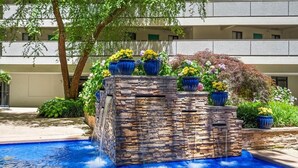 A pool with a stone waterfall feature, surrounded by potted plants and a building in the background. - Direct Oceanfront 3 Bedroom Condo with Outdoor, Indoor, and Kiddie Pools! Sea Watch #909 (Ocean City)