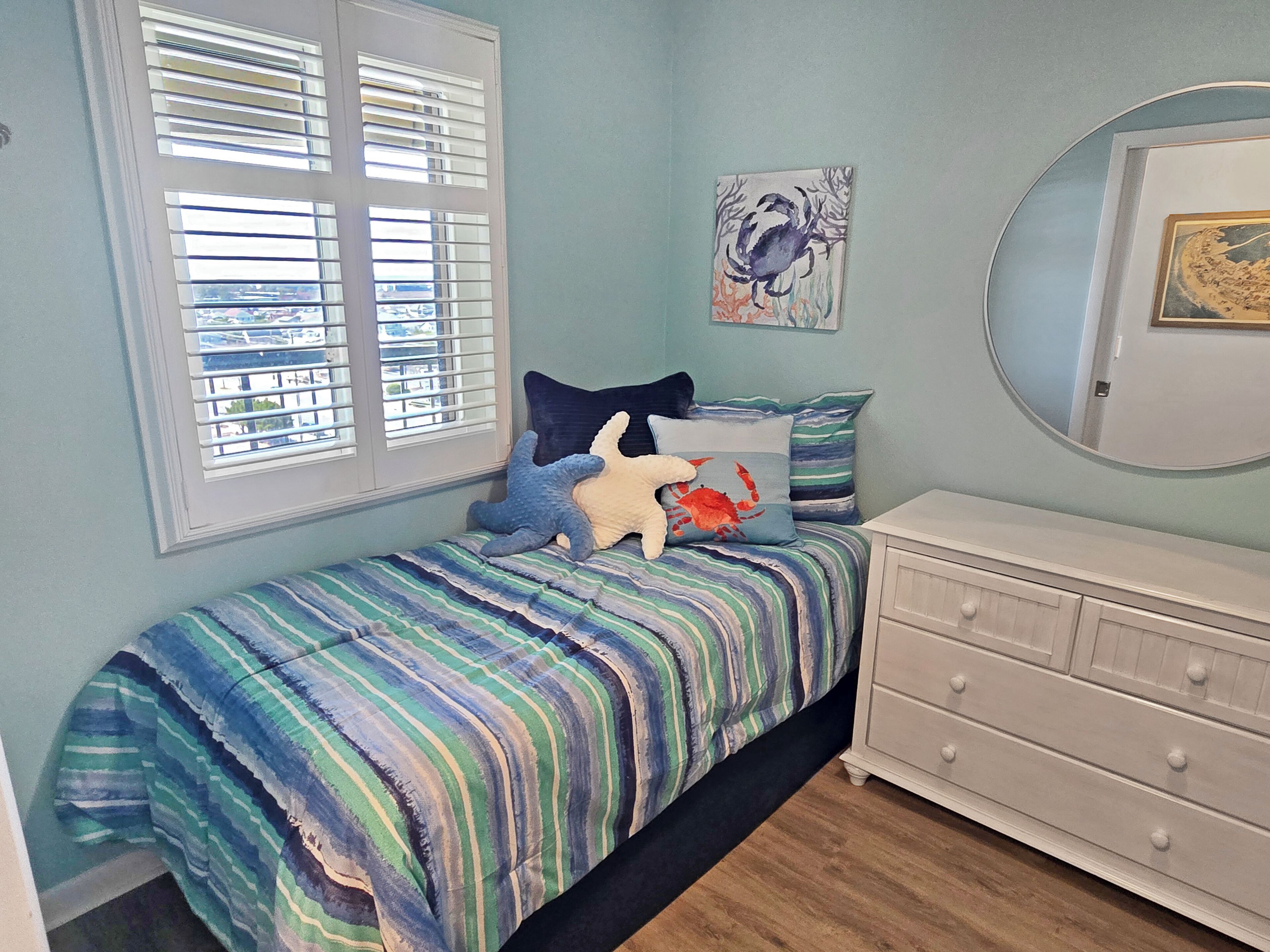 Une chambre à coucher avec un lit, des oreillers, une commode, une fenêtre avec des stores et un mur orné d’un tableau encadré.