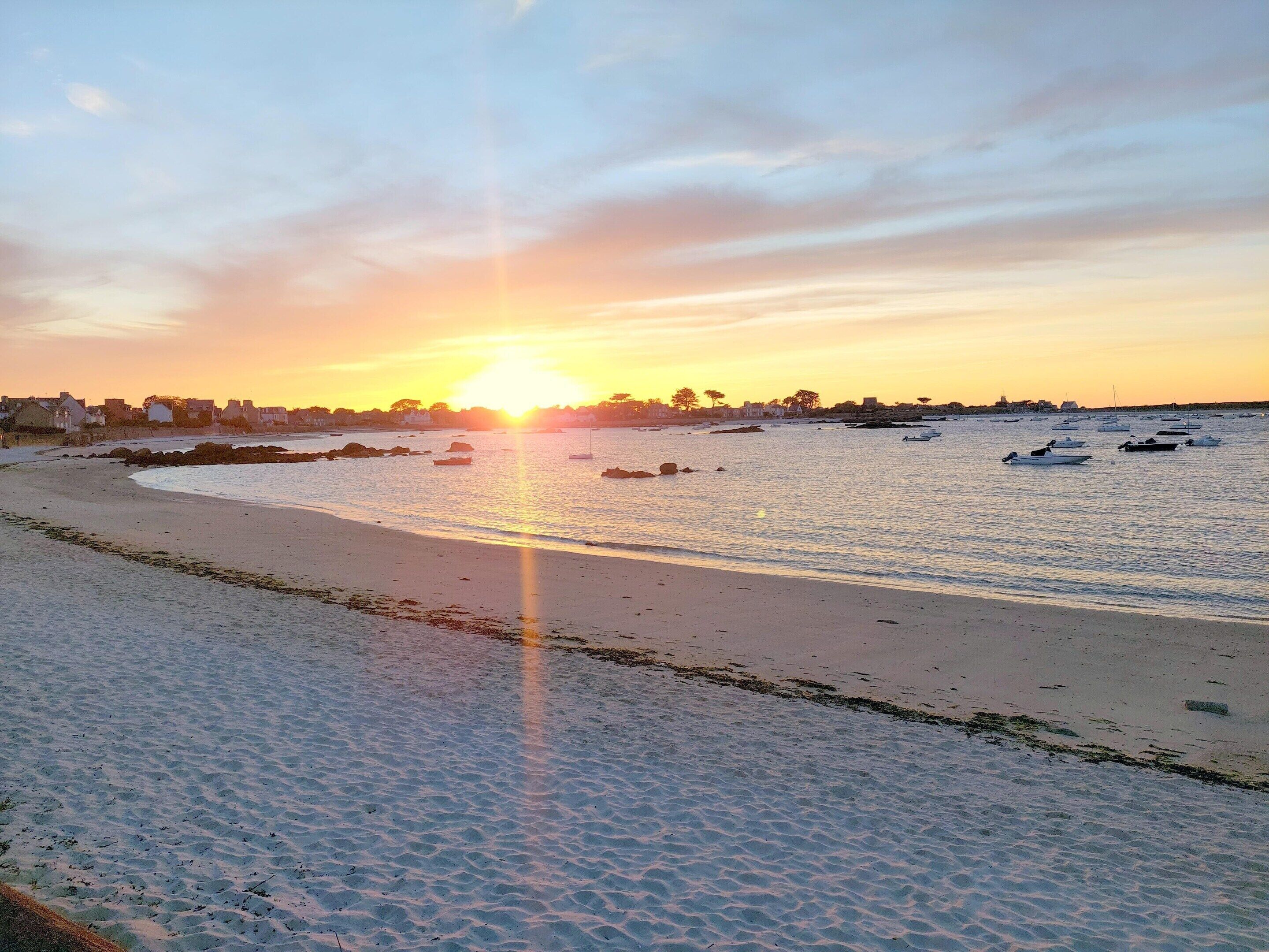Una playa con barcos, un atardecer y una costa lejana con edificios.