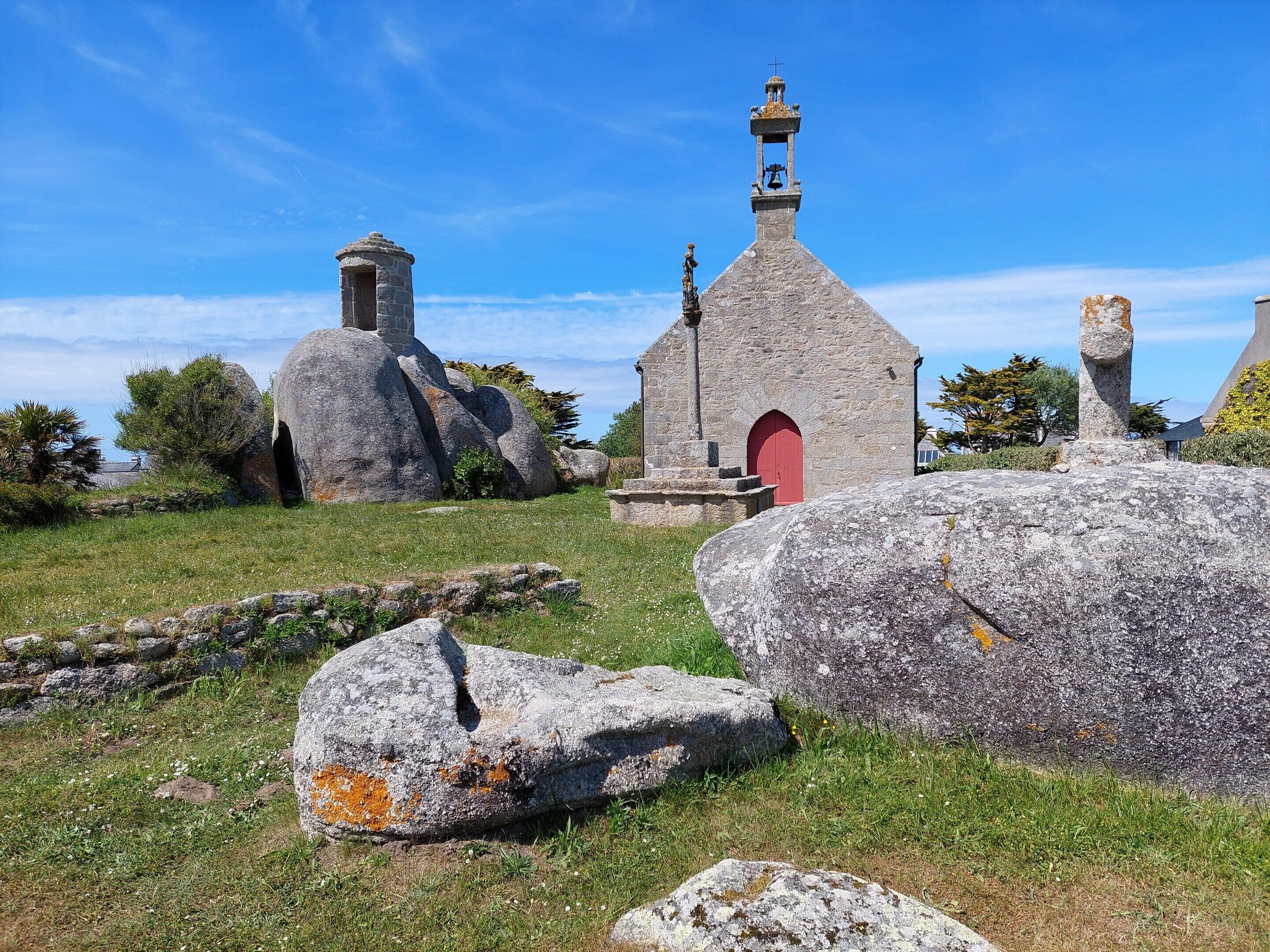 Une église en pierre dotée d’un clocher et d’une porte rouge, entourée de gros rochers et de verdure.
