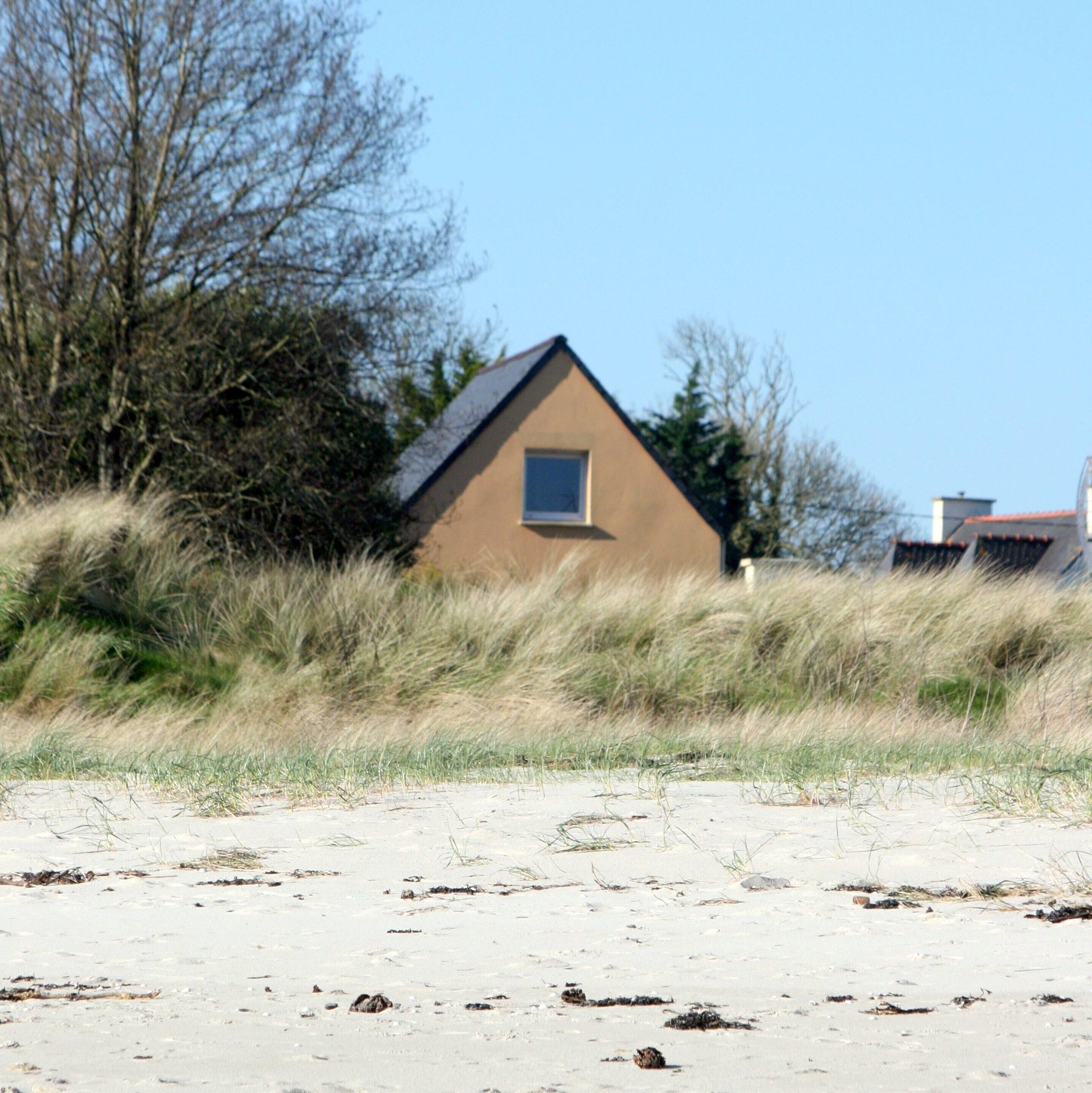 A small, single-story house with a pitched roof, surrounded by sand dunes and sparse vegetation.