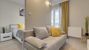 A modern living room with a sofa, a bedside table, a radiator, and a window with curtains. - Mostek 246 - Grey by Grand Apartments (Gdańsk)