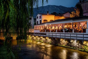 Ein Restaurant am Flussufer mit Außenbestuhlung, Steinmauern und einer Brücke.