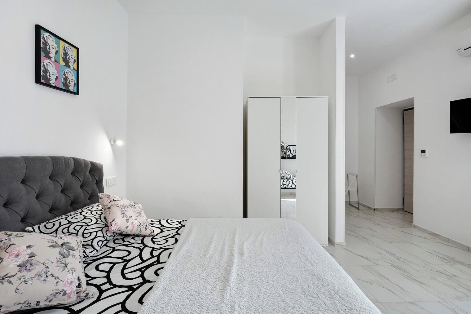 A bedroom with a tufted headboard, a bed with patterned bedding, a white closet, and a TV mounted on the wall.