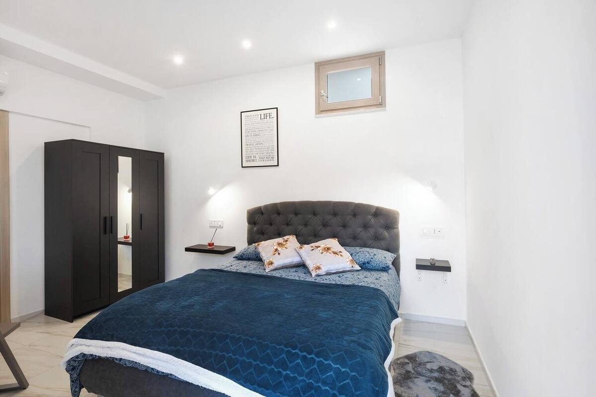 Une chambre à coucher comprenant un lit, une armoire, une table de chevet, une commode, une lampe fixée au mur et une petite fenêtre.