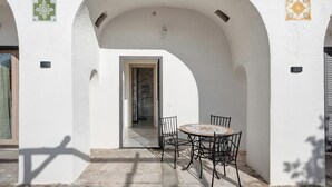 En udendørs patio med et rundt bord og stole, en buegang og en dør der fører til en gang.