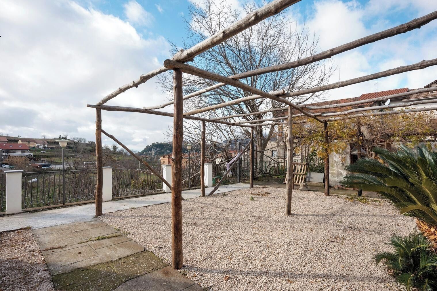 Uma estrutura de pérgula de madeira num pátio com chão de cascalho, rodeada por edifícios e uma vedação.