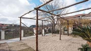 Uma estrutura de pérgula de madeira num pátio com chão de cascalho, rodeada por edifícios e uma vedação.