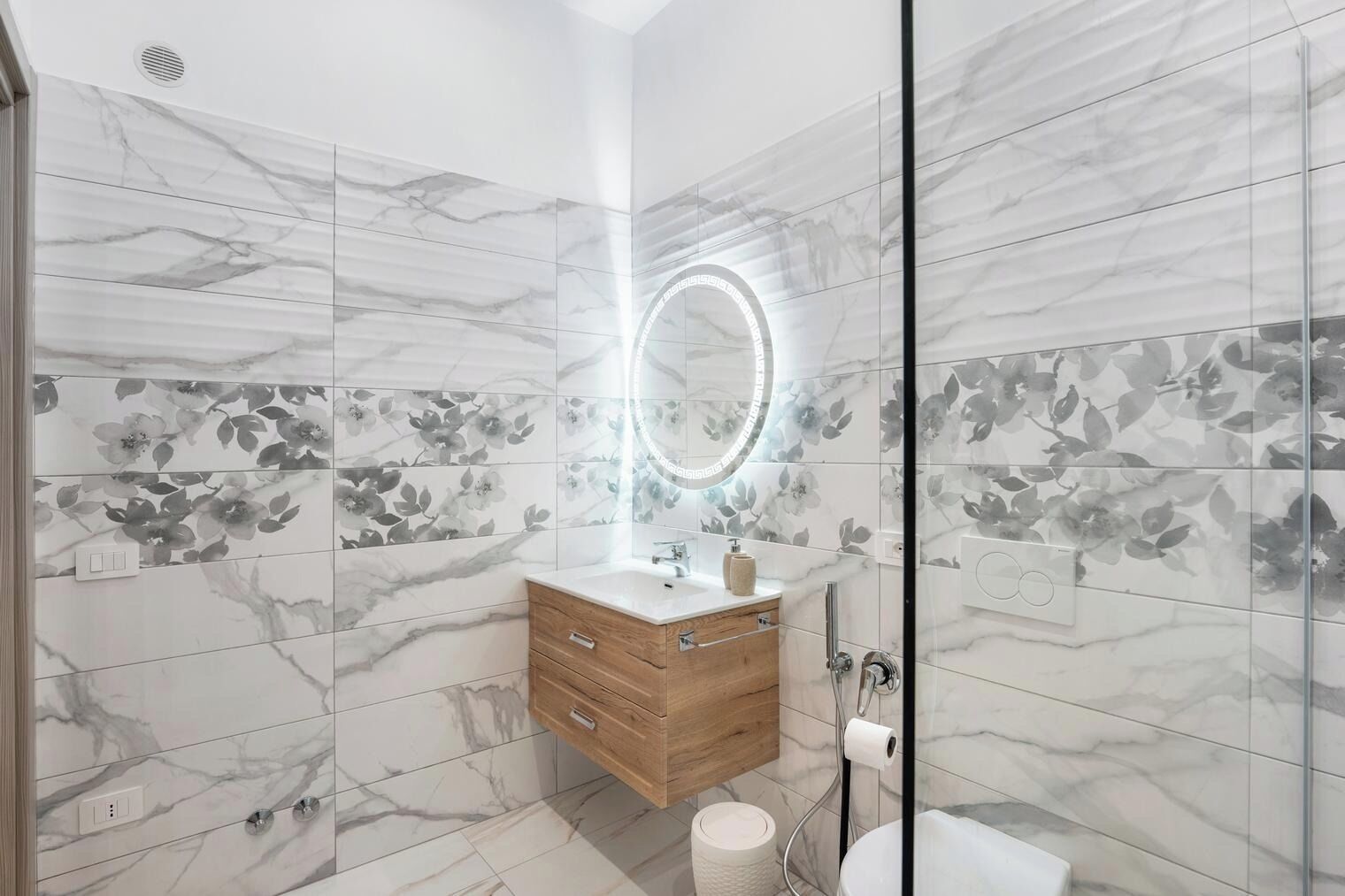 Un baño moderno con baldosas de mármol, tocador de madera, cabina de ducha de cristal y un espejo circular con luces LED.