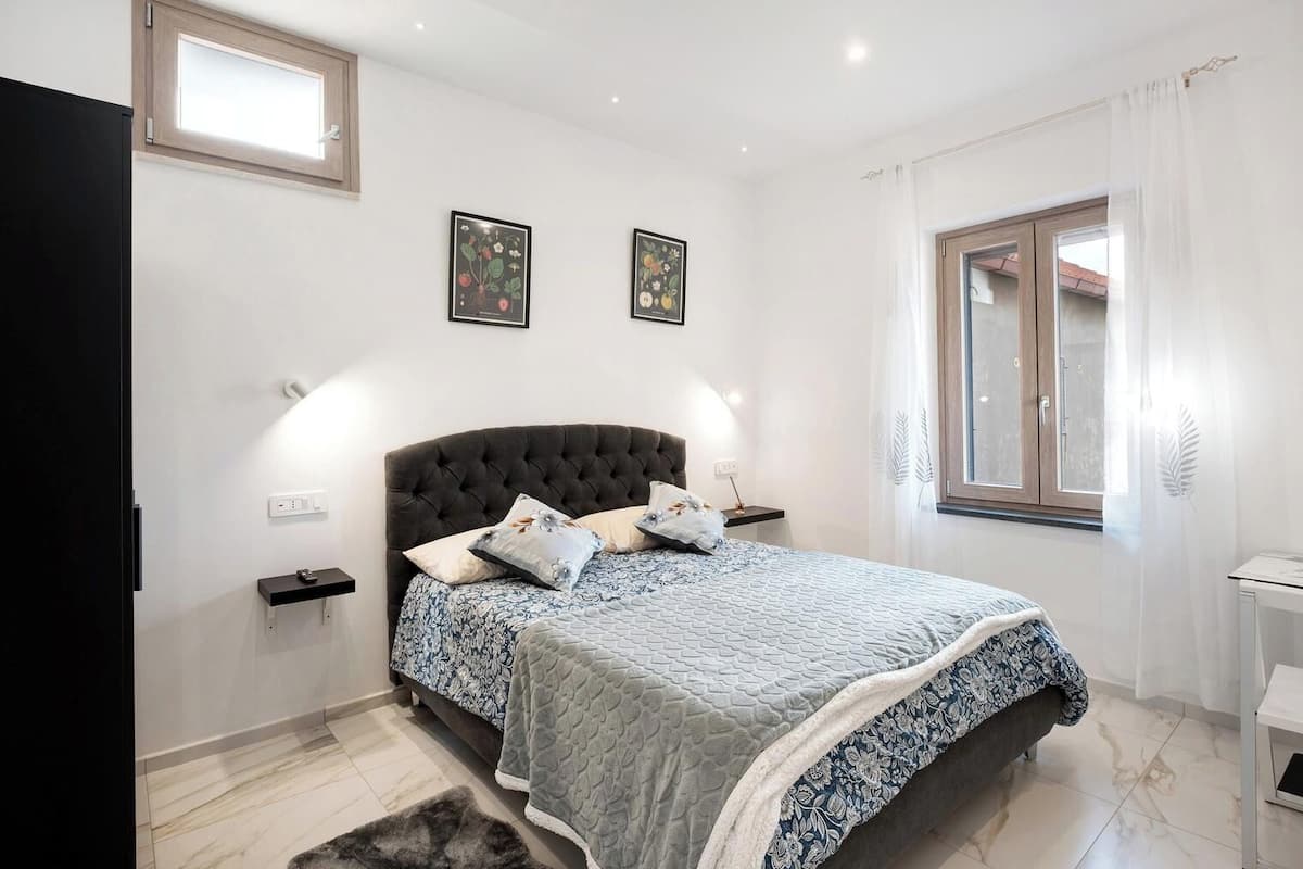 Une chambre à coucher avec une tête de lit capitonnée, deux reproductions botaniques encadrées, une table de chevet avec une lampe et une fen être avec un rideau.