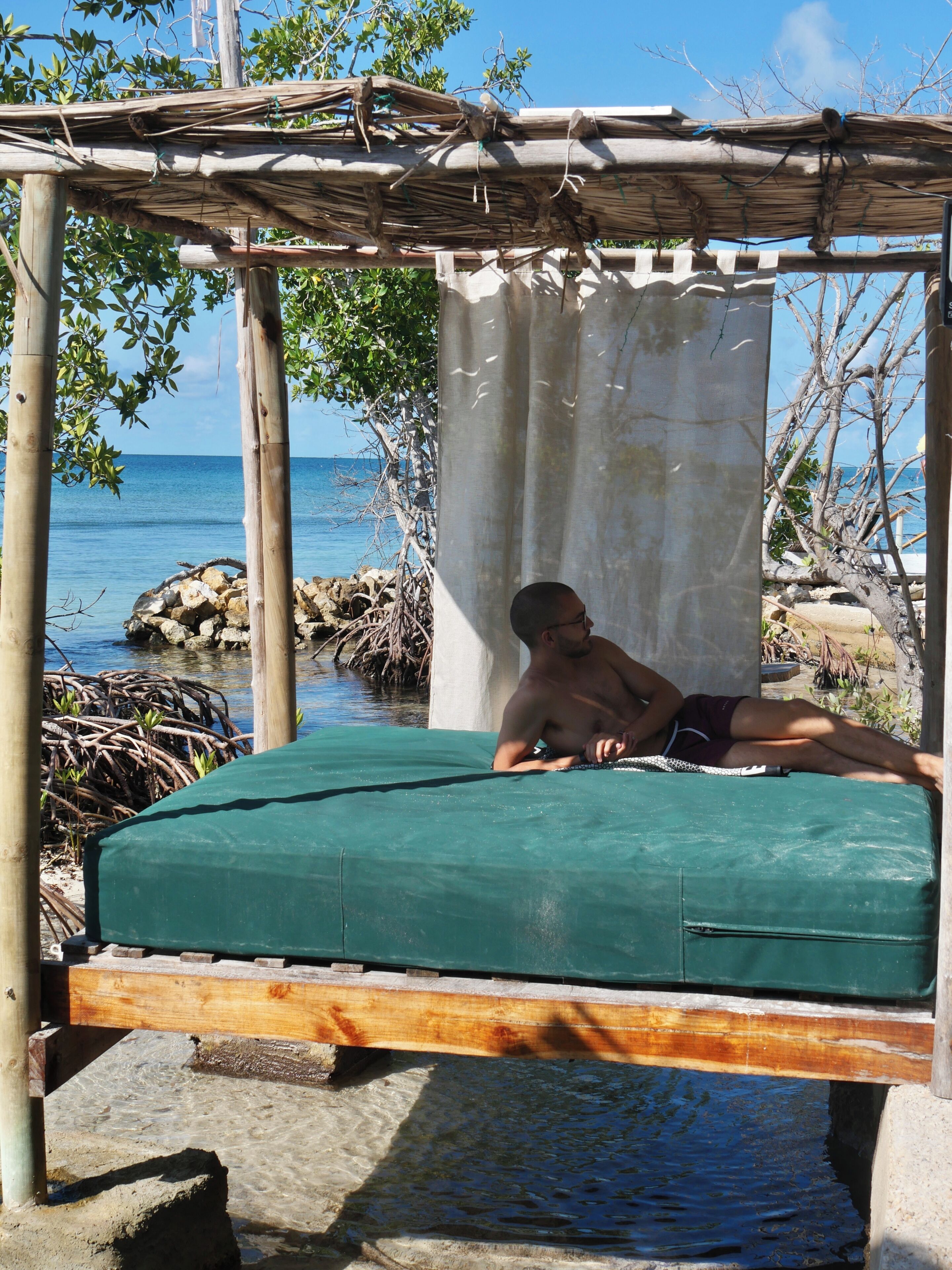 A man lying on a green mat under a wooden structure with a view of the sea.