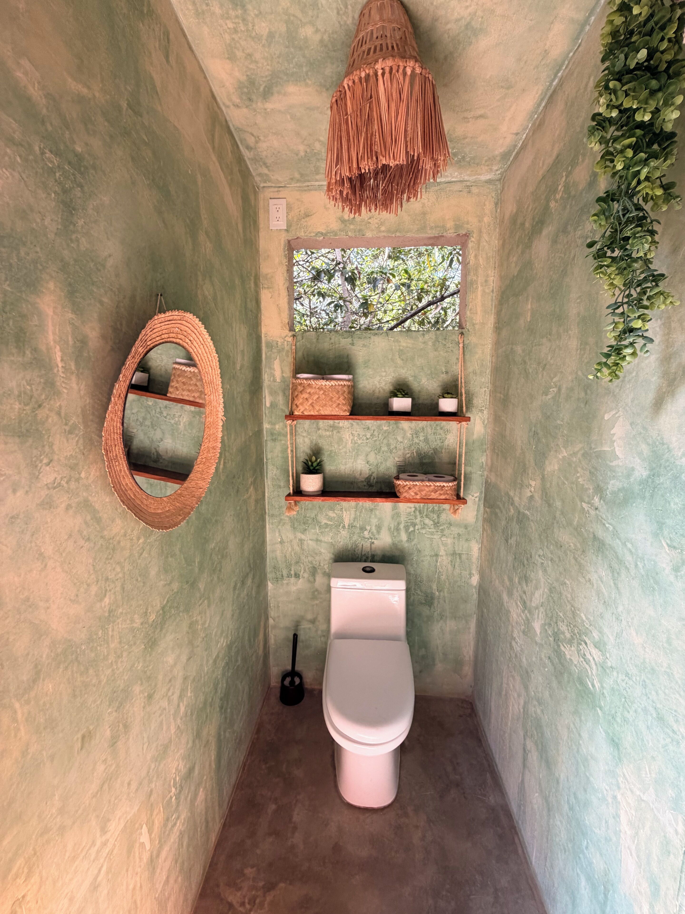 A compact bathroom with a hanging light fixture, a mirror, shelves with plants, and a toilet.