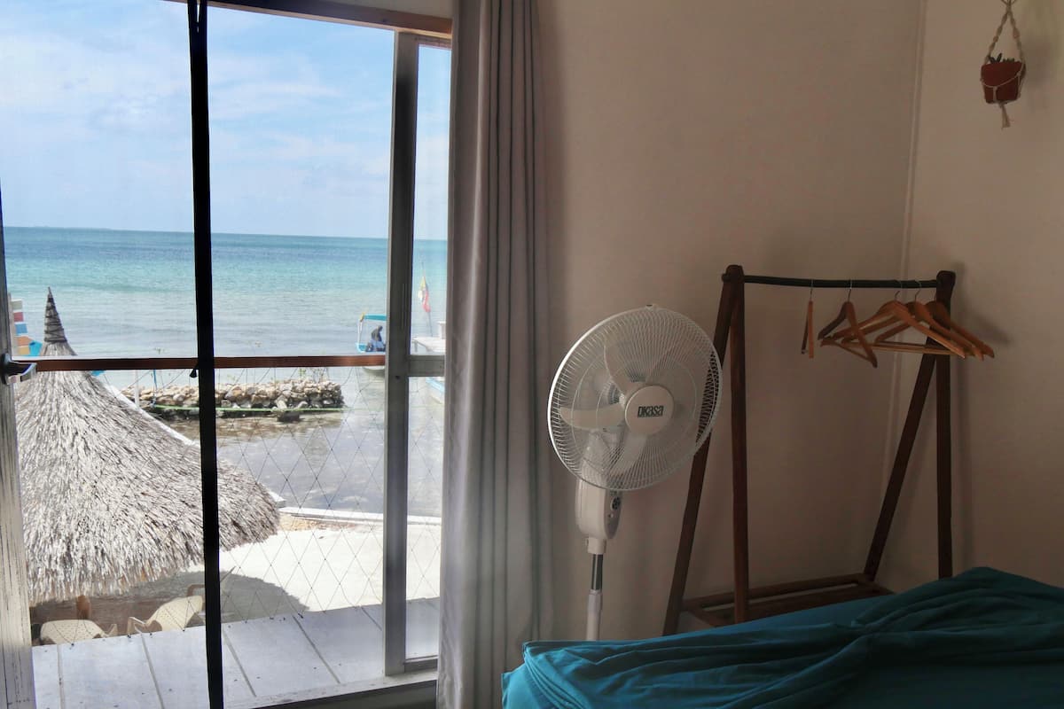 A room with a bed, a standing fan, a clothes rack, and a view of the beach through a large window.