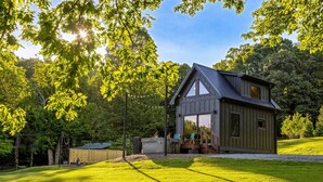 A modern house with a large glass window, surrounded by a well-maintained lawn and trees. - Hot Tub Outdoor Movies Firepit Mtn Views (Mills River)
