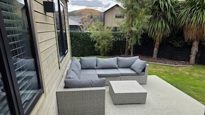 An outdoor seating area with a sofa and two square ottomans, a glass door, and a view of a house and trees. - Near new 4 bed 2 Bath House in Hanleys Farm, Queenstown (Jacks Point)