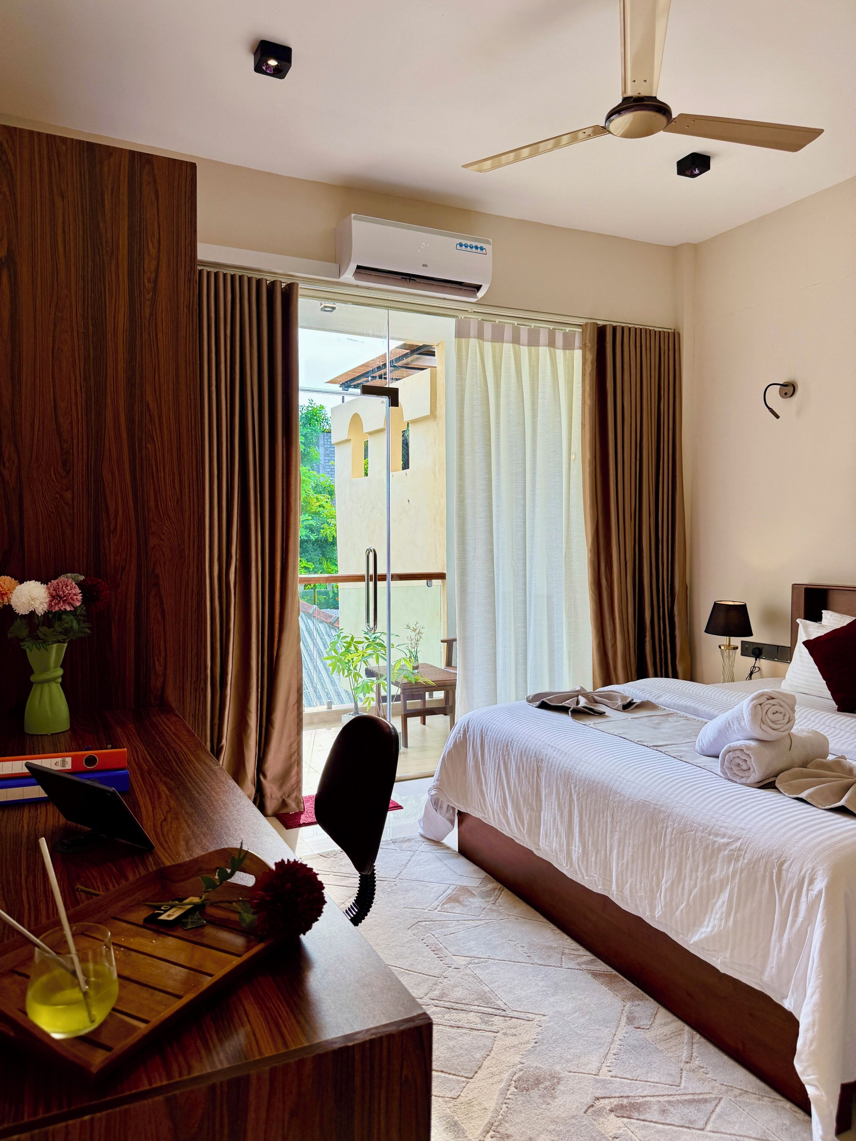 Una habitación de hotel con una cama, un escritorio con una laptop, una silla, una mesa con un vaso de agua, un florero con flores y vistas al exterior.