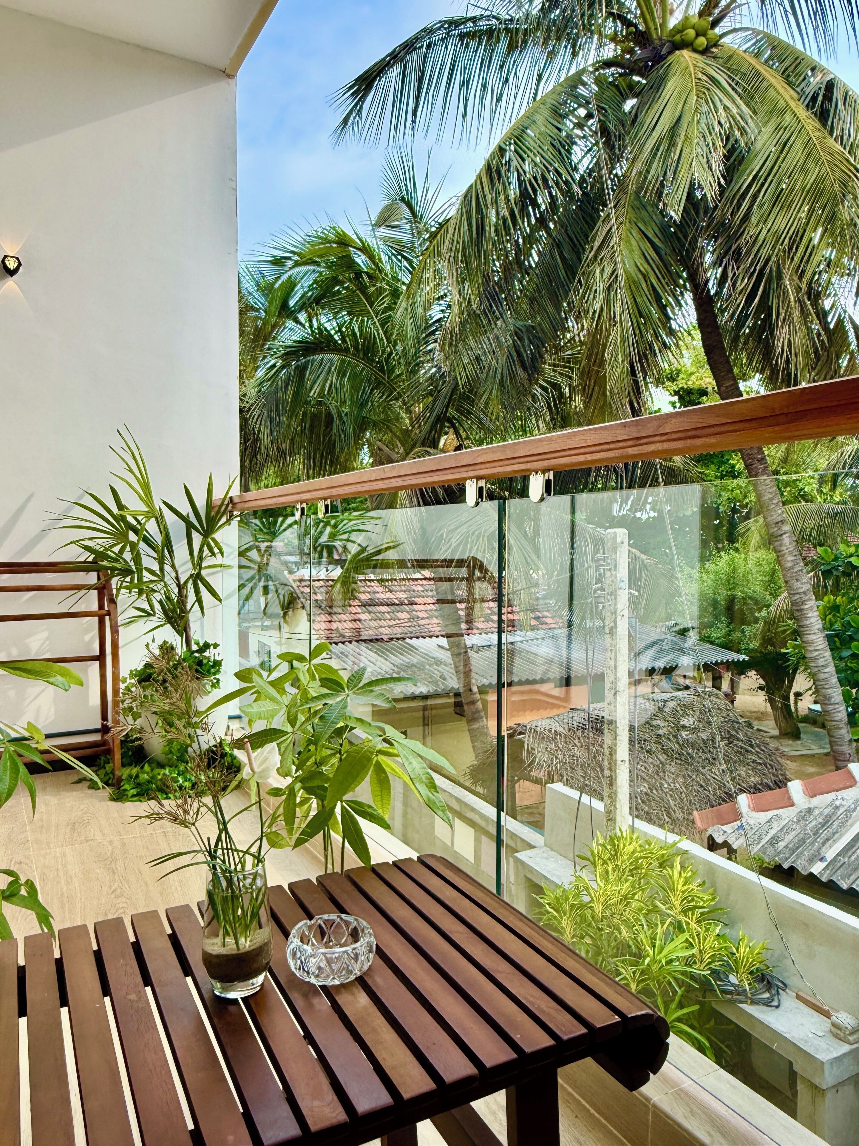 Ein Balkon mit Holztisch, Topfpflanzen und Blick auf Palmen und ein traditionelles Haus.