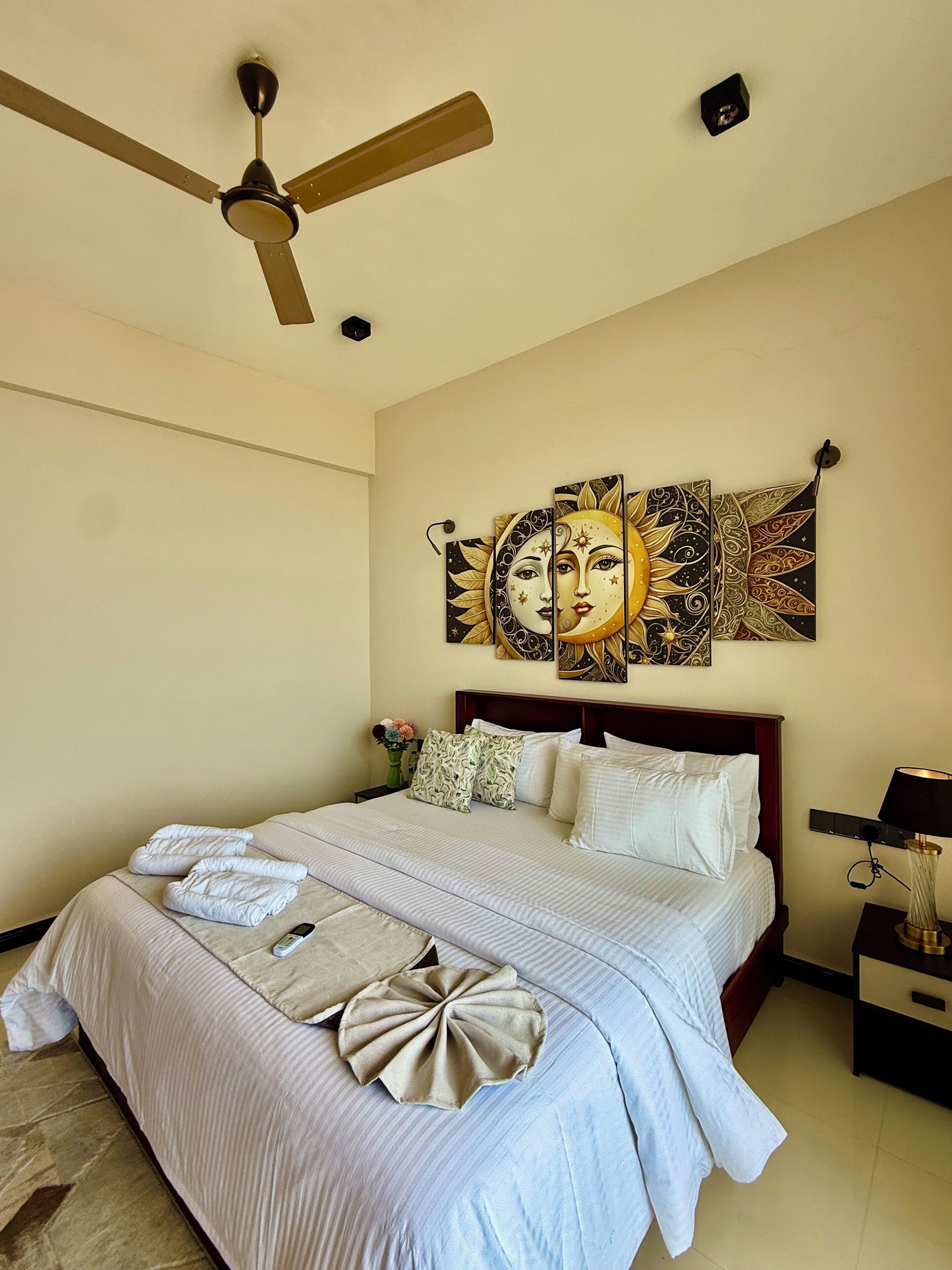 A neatly made bed with white linens, a ceiling fan, and a wall with a large decorative piece.