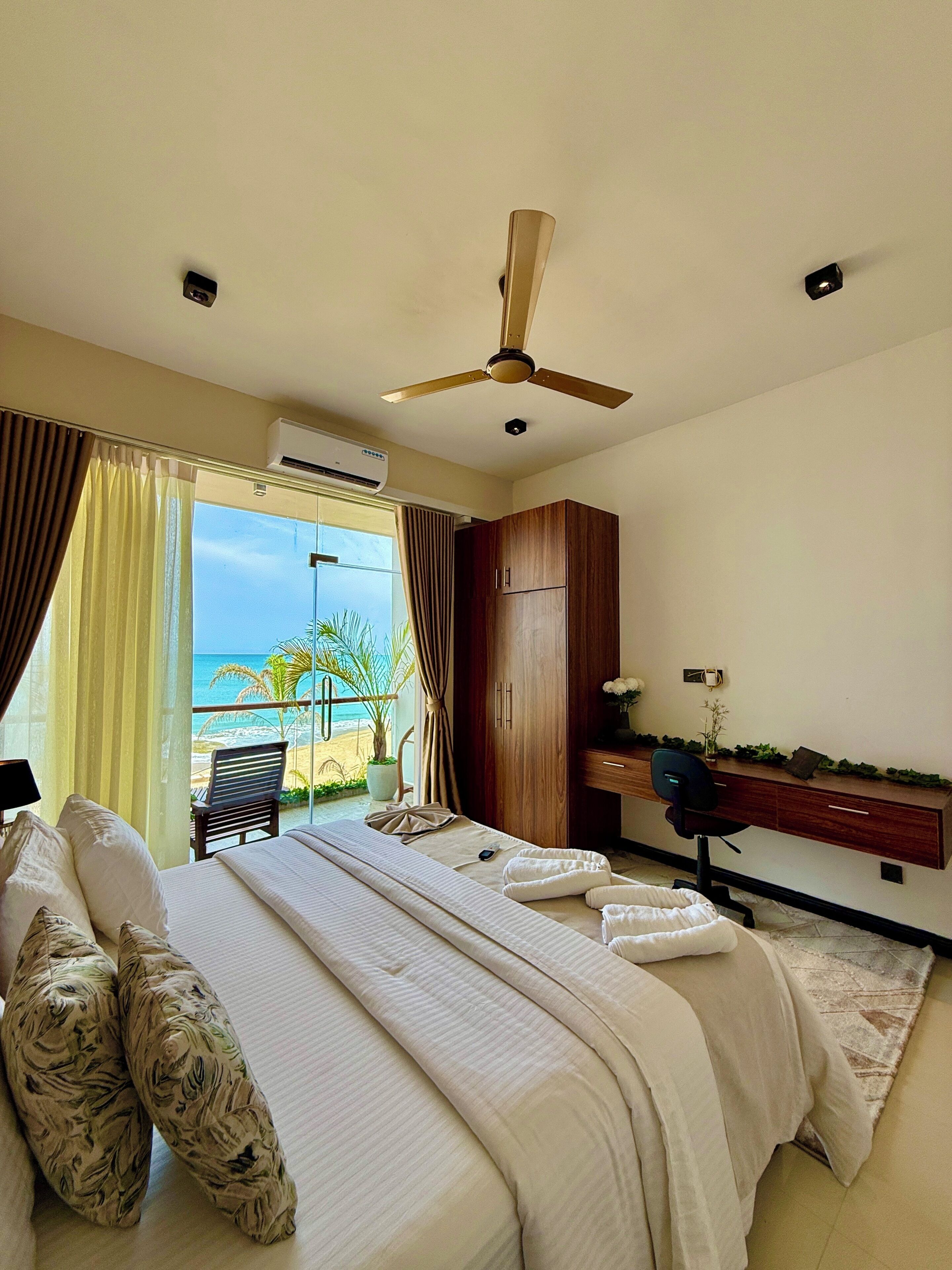 A bedroom with a bed, a desk, a chair, and a view of the beach.