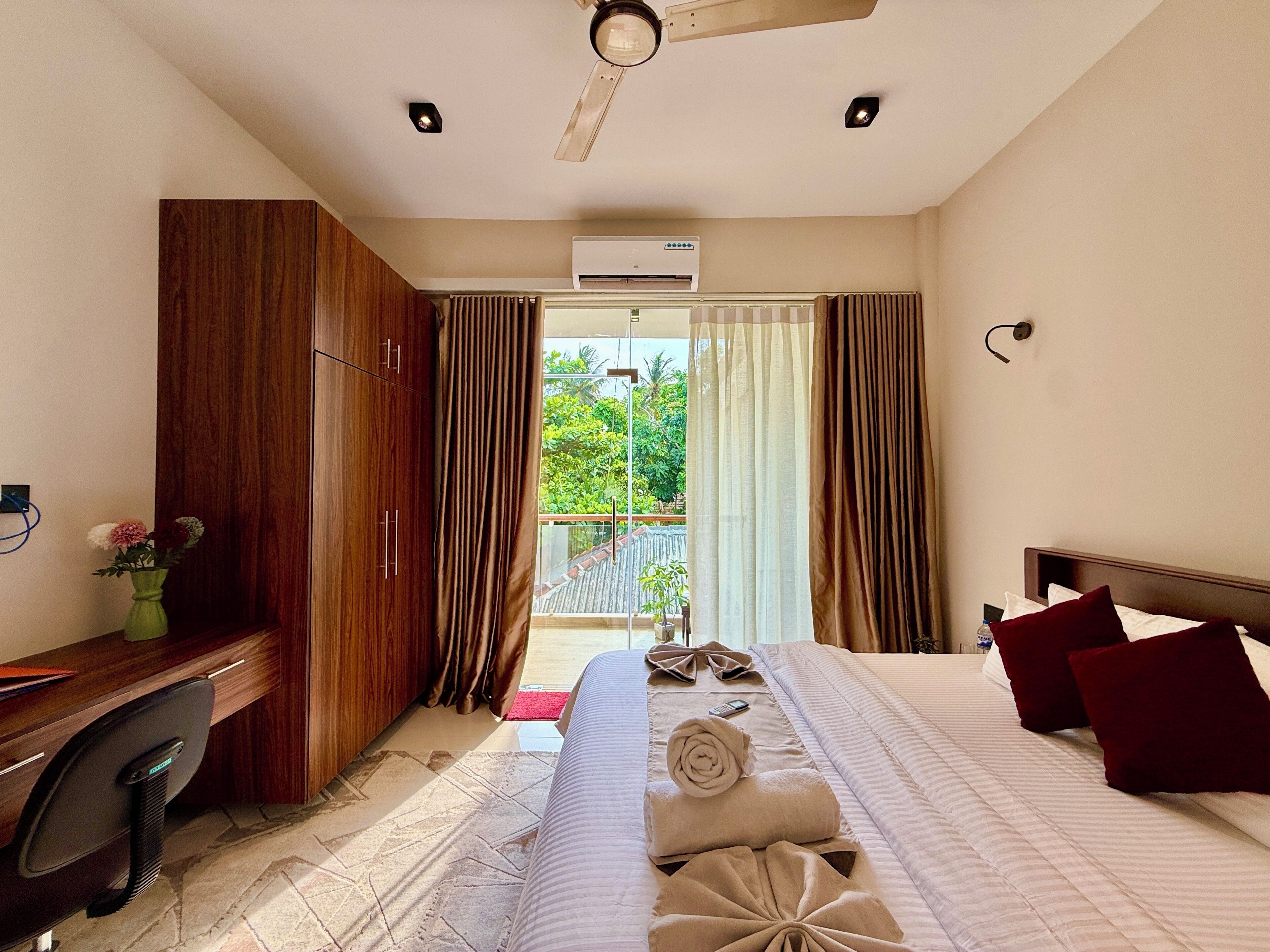 A hotel room with a bed, a desk, a chair, a ceiling fan, an air conditioning unit, and a view of greenery outside.