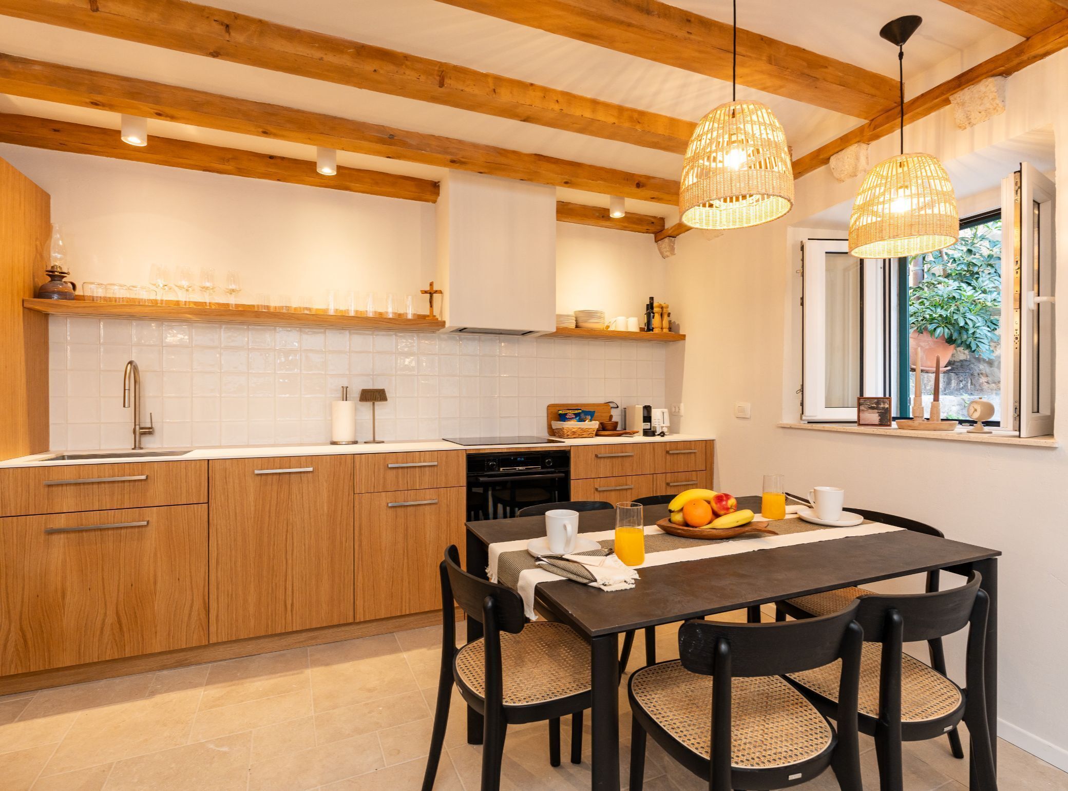 A modern kitchen with wooden cabinets, a dining table with chairs, and a window with a view of greenery.