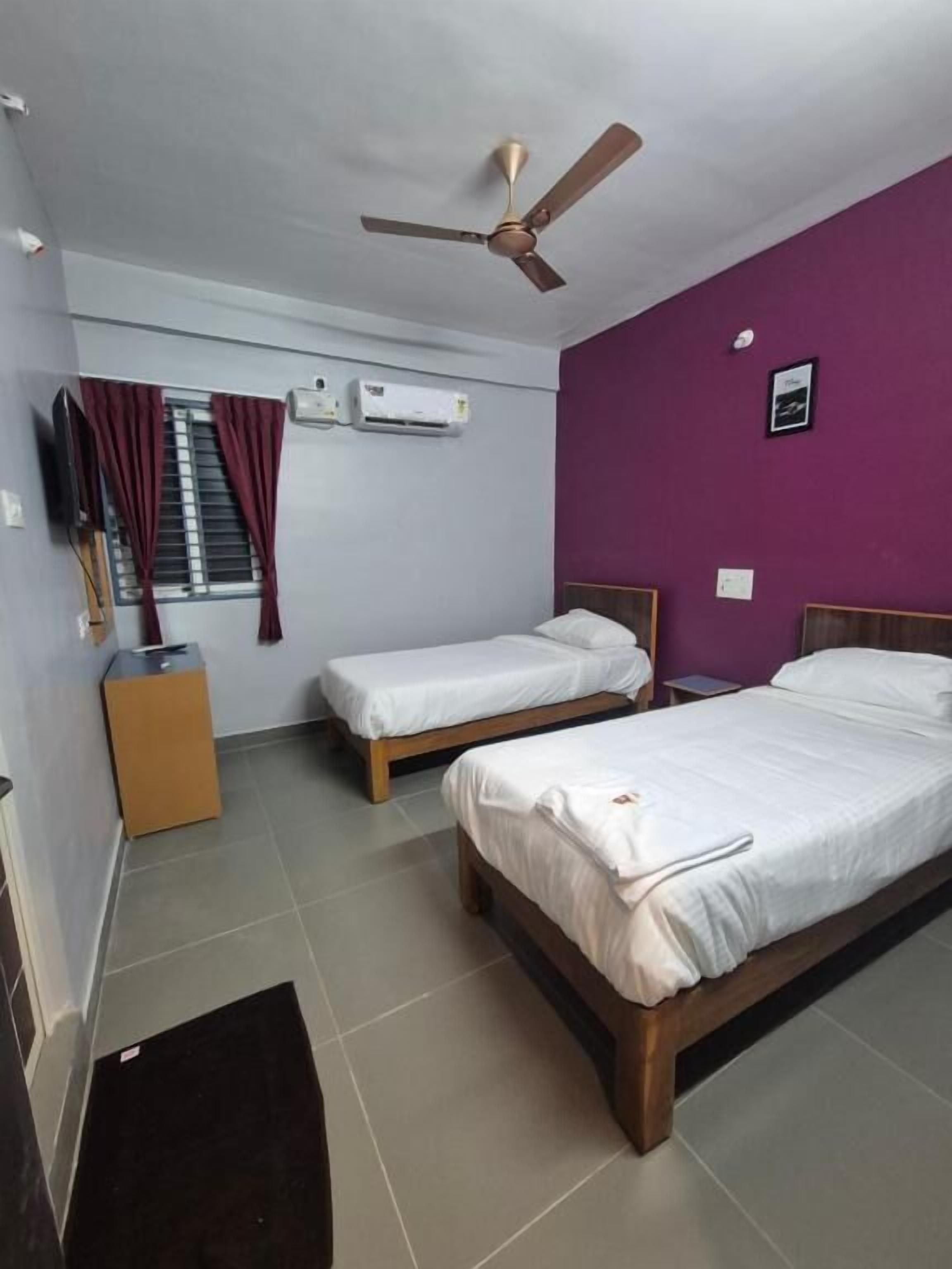 A hotel room with two beds, a ceiling fan, an air conditioning unit, a window with curtains, and a small table.