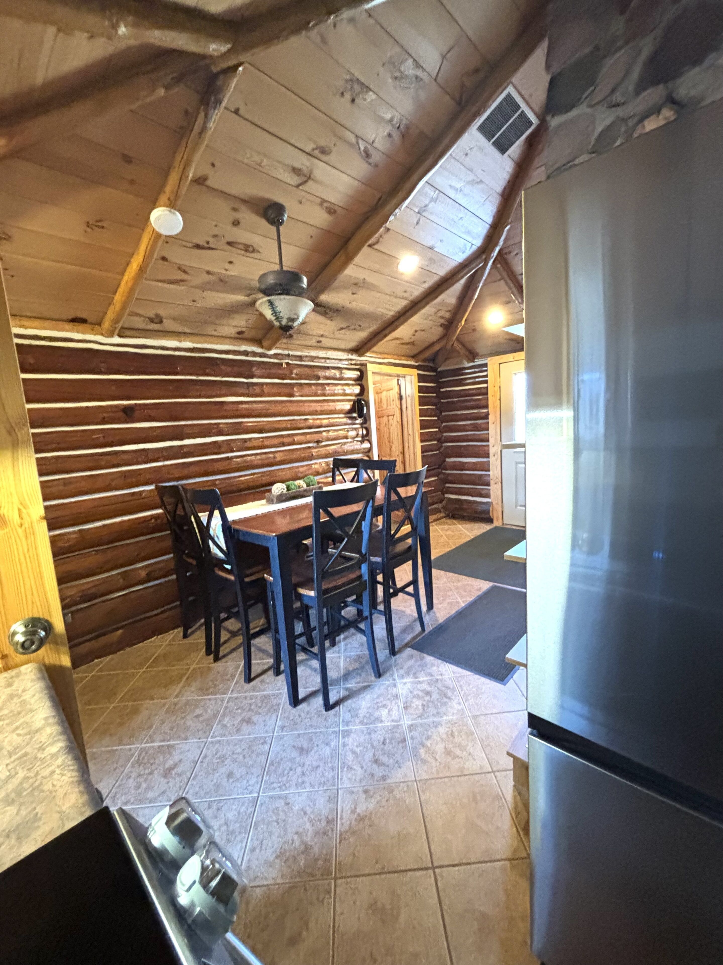 L’intérieur d’une cabane en rondins, avec une table à manger, des chaises, un réfrigérateur et un ventilateur de plafond.