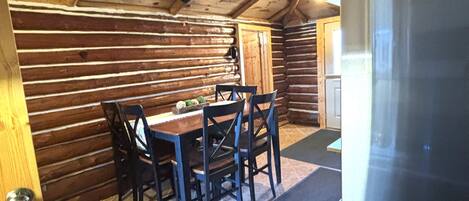 L’intérieur d’une cabane en rondins, avec une table à manger, des chaises, un réfrigérateur et un ventilateur de plafond.