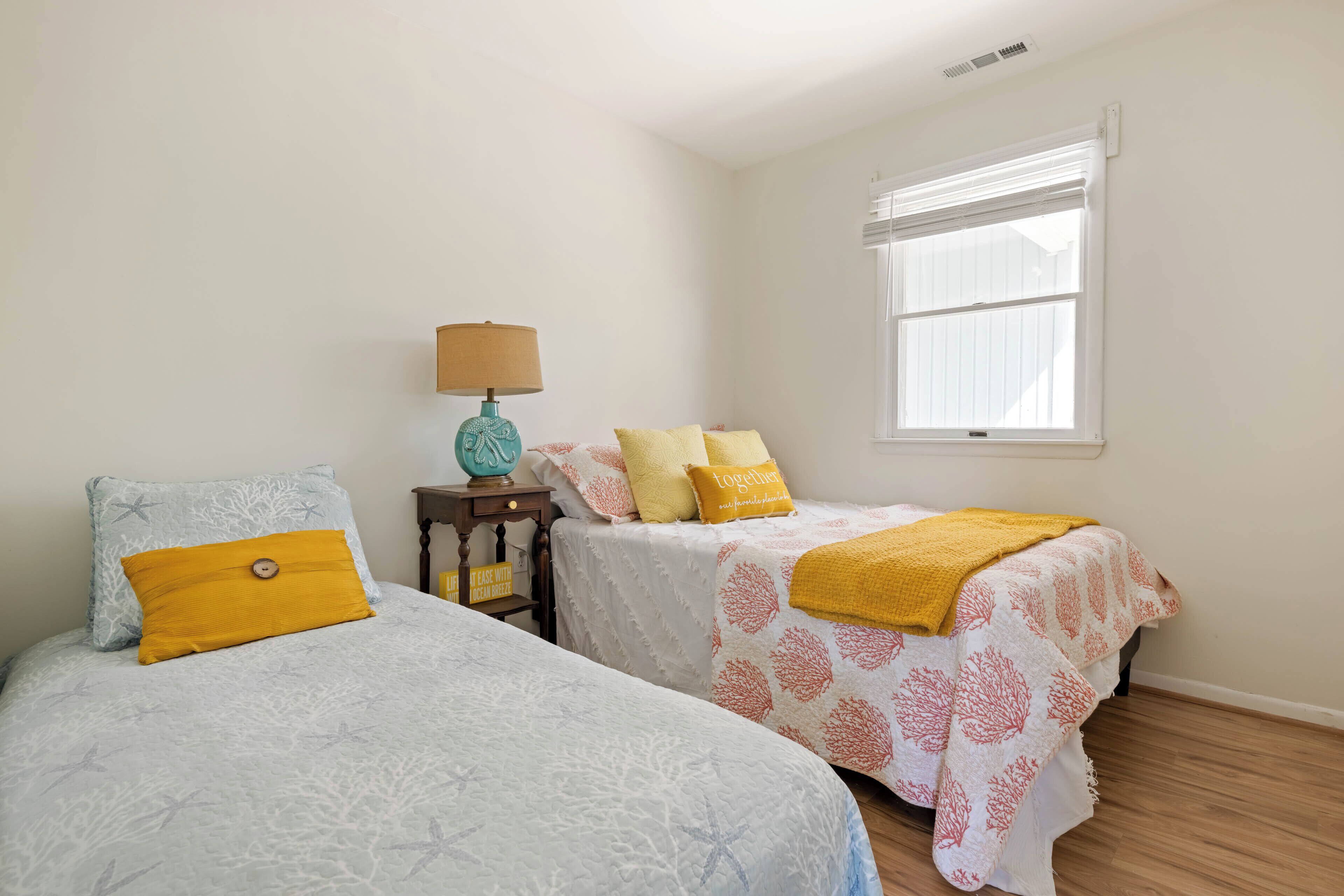 A bedroom with two beds, a nightstand with a lamp, a window with blinds, and a small shelf with a sign.