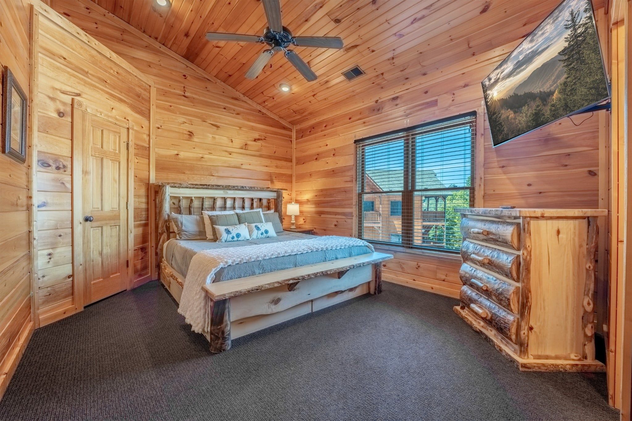 A wooden cabin bedroom with a bed, a ceiling fan, a window with blinds, and a log cabinet.