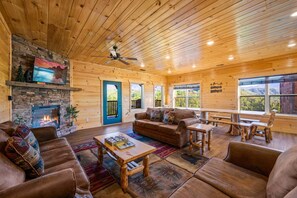 A cozy cabin interior with a stone fireplace, wooden walls, a sofa, a coffee table, and a dining table. - Gatlinburg Views Lodge 5 Bedroom Cabin (Gatlinburg)