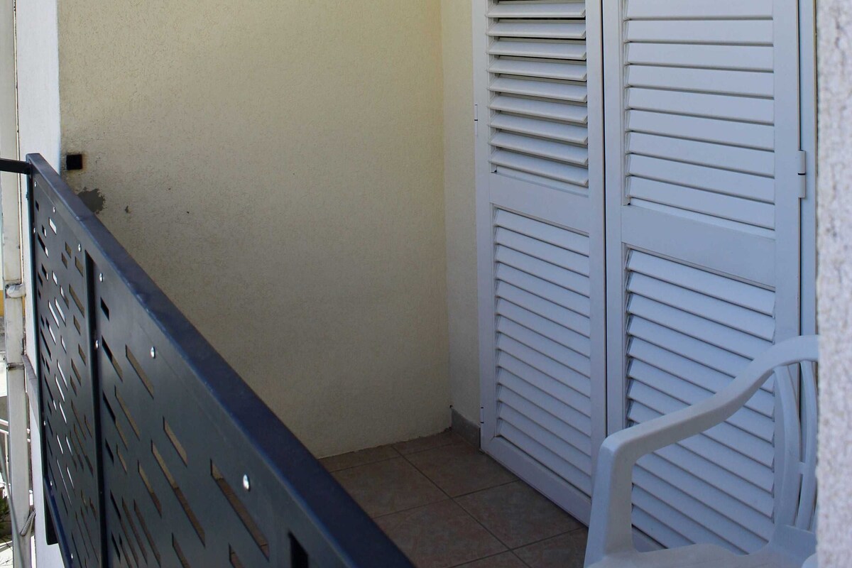 Un balcón con barandilla de metal, una silla de plástico blanco, un ventanal con marco decorativo y un conjunto de puertas blancas con persianas.