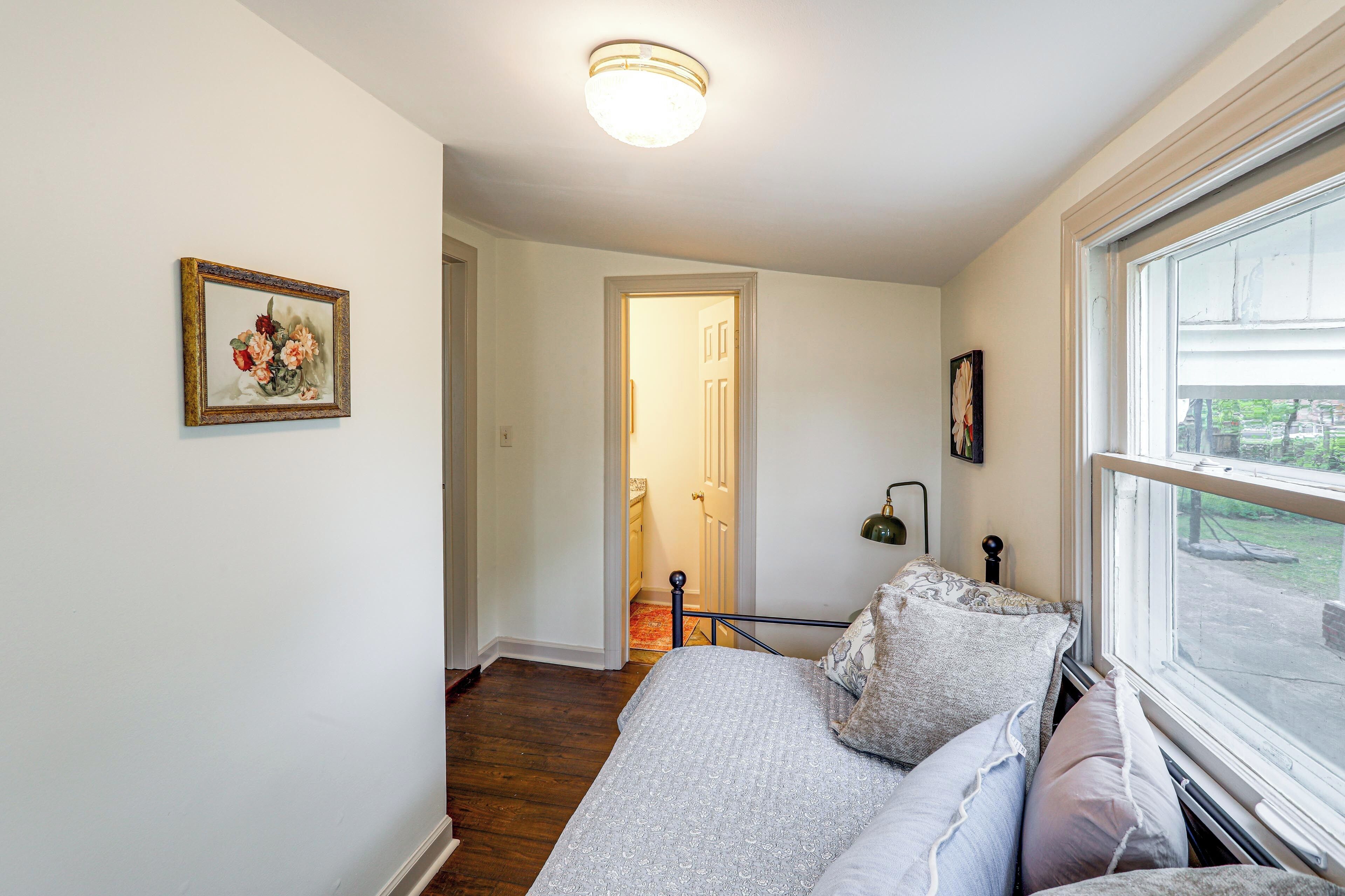 A bedroom with a bed, a window, a door leading to another room, and a wall-mounted picture.