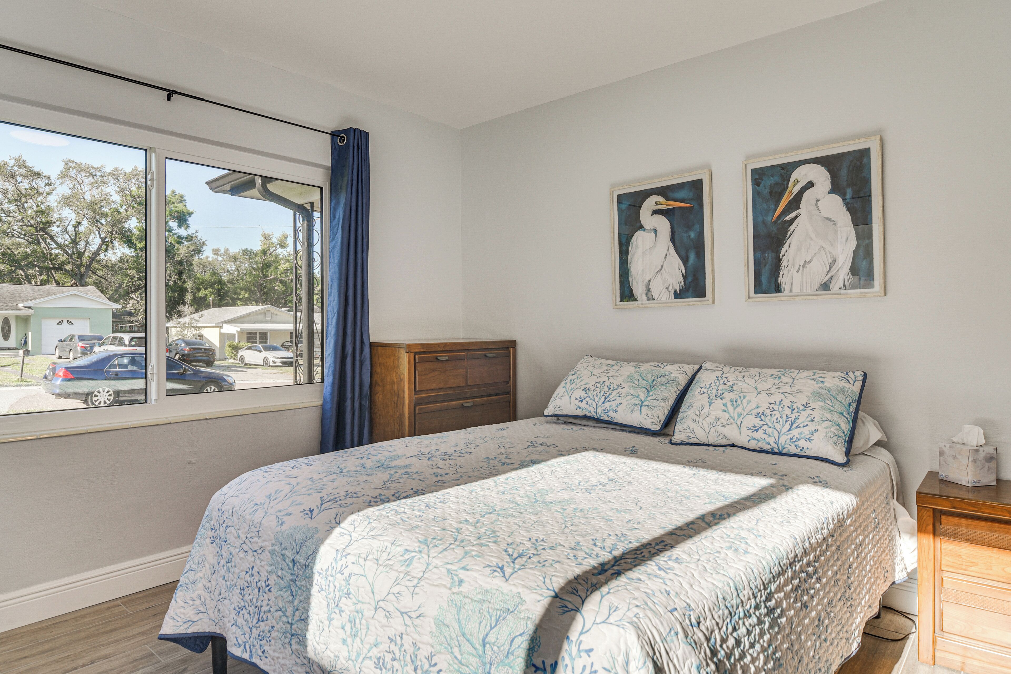 Un dormitorio con cama, cómoda y ventana con vistas a casas y coches.
