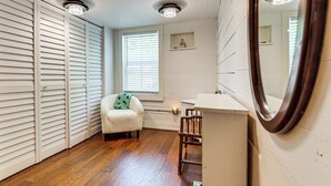 Een kamer met houten vloer, een witte bank, een houten stoel, een witte kaptafel, een spiegel en een raam met zonwering.