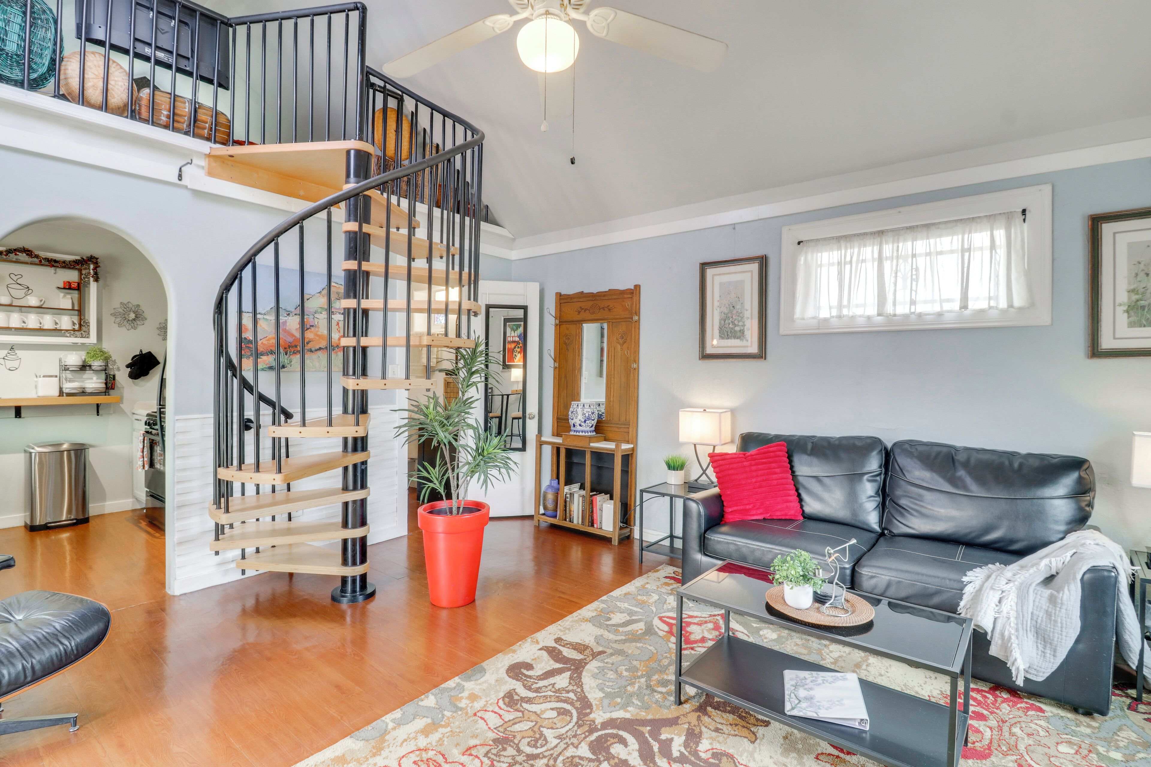 A living room with a black leather sofa, a coffee table, a red throw pillow, a staircase with a wooden railing, and a large potted plant.
